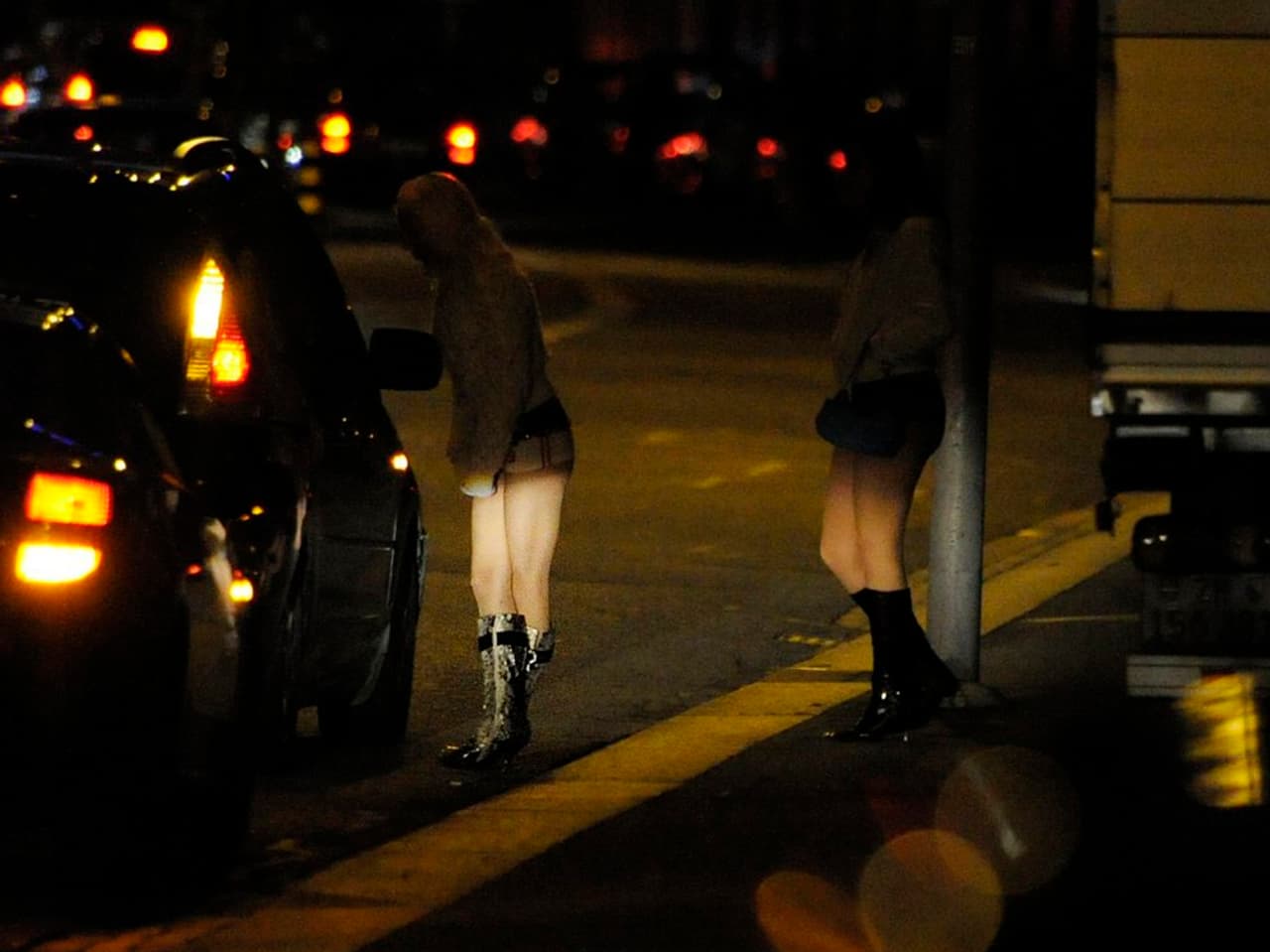 Zwei Prostituierte in hohen Stiefeln stehen im Dunkeln an der Strasse. 