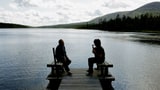 Zwei Männer sitzen auf einem Steg an einem See.