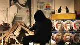 Banksy sitzt in seinem Atelier, Spraykunst im Hintergrund. Er selber ist nur als schwarzer Umriss erkennbar.