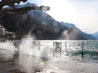 Ufer eines Sees, Wellen peitschen an das Ufer und die Gischt spritzt meterhoch in den Himmel. 