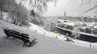 Im Vordergrund steht eine Sitzbank mit etwa 20 Zentimeter Schnee darauf. Die Bank schaut Richtung Bildhintergrund. Zu sehen ist eine Fluss und die Stadt mit weissen Dächern.