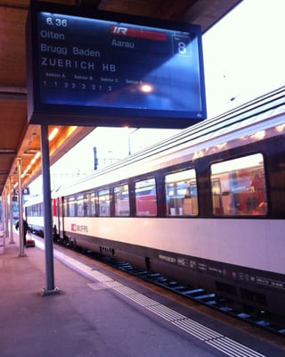 Ein Zug fährt am Bahnhof ein. Auf einer Tafel wird angezeigt, dass der Zug um 6 Uhr 36 Minuten nach Aarau fährt.