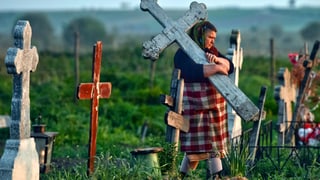 Eien Frau auf einem ländlichen Friedhof trägt einen steinernen Grabstein in Kreuzform.