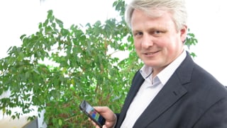 Ein Mann steht vor einer Büropflanze und hält ein Smartphone in der Hand.