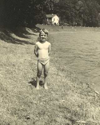 Schwarz-Weiss-Fotografie mit einem kleinen Bub, der in Badehosen am Ufer eines Flusses steht.