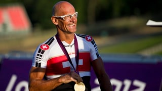Ein Mann mit Glatze und Brille zeigt stolz eine Goldmedaille.