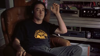 John Cusack sitzt auf einem Sessel. Er trägt Kopfhörer. John Cusack sitzt auf einem Sessel. Er trägt Kopfhörer.