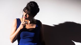 Noemi Nadelmann in einem blauen und kurzen Kleid. Sie lehnt an eine Wand und raucht.
