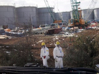 Im Hintergrund mehrere Reaktoren, im Vordergrund zwei Arbeiter in weissen Schutzanzügen mit gelben Helmen
