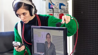 Eine Frau mit Kopfhörer und Mikrofon steht hinter einem Bildschirm.
