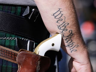 Nahaufnahme: Ein Mann trägt einen Pistolengurt mit Pistolengriff, auf seinem Arm ist das Tattoo "We the People" zu sehen.