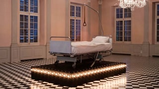 Ein Spitalbett auf einem Sockel mit Lichtern in einem Saal mit Kronleuchtern. Ein Spitalbett auf einem Sockel mit Lichtern in einem Saal mit Kronleuchtern.