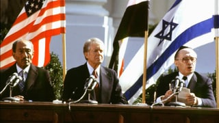 Die drei Politiker sitzen vor Landesflaggen.