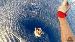 Ein dunkelhäutiger Mann wird mit einer Seilwinde aus einem blauen Ozean in einen Helikopter gehievt. Das Bild wurde aus dem Helikopter geschossen, man sieht nur eine Hand, das Seil und weiter unten ein Mann, der daran hängt und nach oben in die Kamera blickt. Unter ihm das blaue Meer.