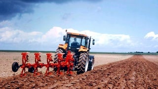Ein Traktor mit rotem Pflug zieht Furchen in eine rotbräunliches Feld. Darüber ein hellblauer Himmel mit weissen Wolken.