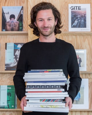 Ein junger Mann posiert mit einem Stapel Zeitschriften in den Händen vor einem Bücherregal.