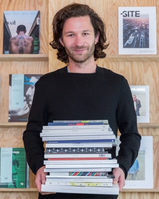 Ein junger Mann posiert mit einem Stapel Zeitschriften in den Händen vor einem Bücherregal. Ein junger Mann posiert mit einem Stapel Zeitschriften in den Händen vor einem Bücherregal.