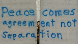 Zwei Jungen schauen hinter einer Mauer hervor auf der steht: "Peace comes agreement not Separation"