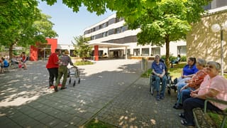 Gebäude im Hintergrund, davor Menschen am Rollator und im Rollstuhl. Gebäude im Hintergrund, davor Menschen am Rollator und im Rollstuhl.