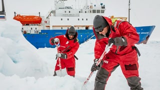 Teilnehmer der Expedition räumen einen Landeplatz für den Helikopter frei. Unmengen von Schnee umgeben sie, sie tragen rote Anzüge. 