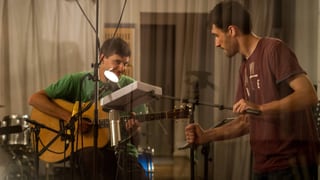 Ein junger Mann mit Gitarre sitzt in einem Tonstudio, ein zweiter junger Mann hält das Mikrofon.