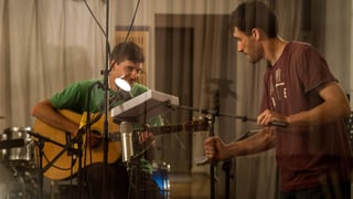 Ein junger Mann mit Gitarre sitzt in einem Tonstudio, ein zweiter junger Mann hält das Mikrofon.