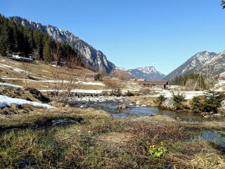 Ein Bach in der Mitte sammelt das Wasser des schmelzenden Schnees, im Hintergrund Berge und eine Hütte.