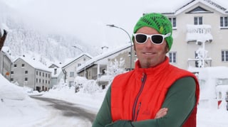 Mann in orangem Gilet, grüner Mütze und weisser Sonnenbrille, dahinter verschneites Dorf