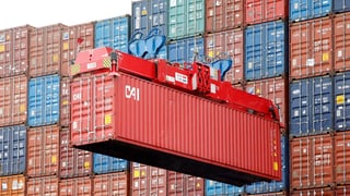 Roter Schiffscontainer hängt an Kranseilen vor einer Wand weiterer Container