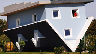 Ein hellblaues Einfamilienhaus auf dem Kopf, das heisst auf dem Giebeldach, auf einer Wiese.