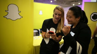 Zwei junge Frauen schauen lachend auf ein Smartphone; an der Wand das Snapchat-Logo.