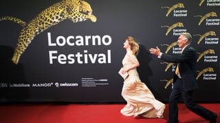 eine Frau in Abendkleid und ein Mann in Anzug rennen über den roten Teppich. Im Hintergund eine schwarze Wand, auf der gross der Schriftzug "Locarno Festival" und ein springender Leopard zu sehen sind.