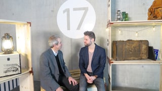 Zwei Herren sitzen auf einer Holzkiste. Über ihren Köpfen die Zahl 17.