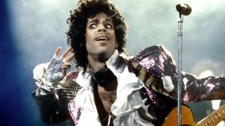 Prince mit rosa Glitzerjacke, Spitzenhandschuhen und Gitarre hält sich lauschend eine Hand ans Ohr.
