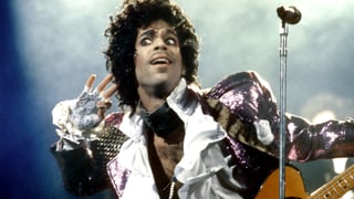 Prince mit rosa Glitzerjacke, Spitzenhandschuhen und Gitarre hält sich lauschend eine Hand ans Ohr. Prince mit rosa Glitzerjacke, Spitzenhandschuhen und Gitarre hält sich lauschend eine Hand ans Ohr.