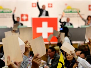 Mann auf einem Podest mit Schweizer Flagge, vorne Hände mit Zetteln.