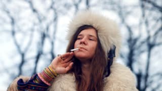 Eine Frau in Hippie-Pelz raucht eine Zigarette, im Hintergrund stehen dunkel entlaubte Bäume.