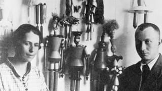 Sophie Tauber und Hans Arp stehen vor einer Wand mit Marionetten. Sophie Tauber und Hans Arp stehen vor einer Wand mit Marionetten.