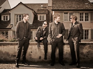 Fotografie in Brauntönen mit den Musikern. Drei stehend, einer sitzen vor, beziehungsweise auf einer Mauer.