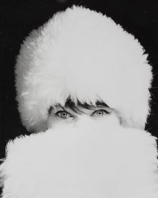 Schwarz-Weiss-Foto: Eine Frau mit weissem Pelzhut verdeckt ihr Gesicht so mit einem weissen Pelz, dass nur noch ihre Augen zu sehen sind.
