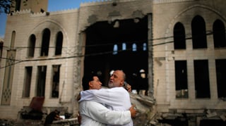 Zwei Männer umarmen sich vor einer zerbombten Moschee.