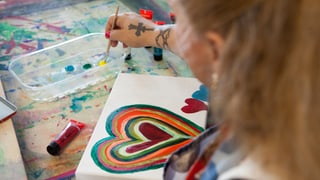 Eine Frau tunkt einen Pinsel in Farbe. Sie sitzt vor einer Leinwand, auf der verschiedene Herzen gemalt sind. 