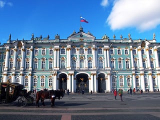 Der Winterpalast unter strahlend blauem Himmel: Vor dem weiss-türkis-goldenen Gebäude steht eine Kutsche.