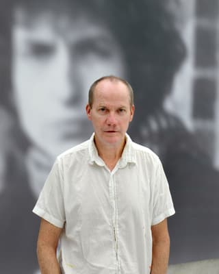 Der Künstler Richard Prince steht in einem Kurzarmhemd vor einem seiner Gemälde.