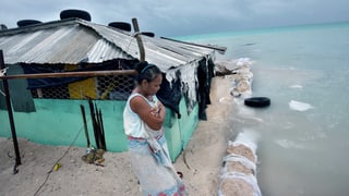 Eine Frau steht auf dem Festland und schaut mit verschränkten Armen auf die Wassermassen des Meeres, die bereits ihr Haus erfasst haben.