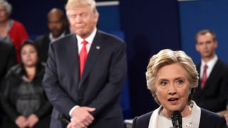 Clinton während der TV-Debatte mit Mikrofon, Trump leicht unscharf hinter ihr