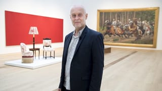Ein Herr im Anzug steht in einem Ausstellungsraum, hinter ihm ein Gemälde und Möbel Ein Herr im Anzug steht in einem Ausstellungsraum, hinter ihm ein Gemälde und Möbel