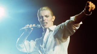 Bowie mit ausgestrecktem linkem Arm und Mikrofon in der rechten HAnd, singend auf einer Bühne.