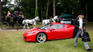 Ein Mann mit Frack und Zylinder geht an einem Ferrari vorbei. Im Hintergrund eine Pferdekutsche.