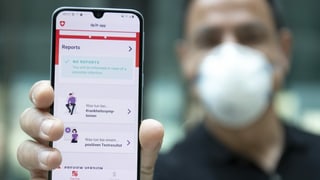 Ein Mann mit Gesichtsmaske hält ein Smartphone in die Kamera, auf der eine App läuft. Ein Mann mit Gesichtsmaske hält ein Smartphone in die Kamera, auf der eine App läuft.